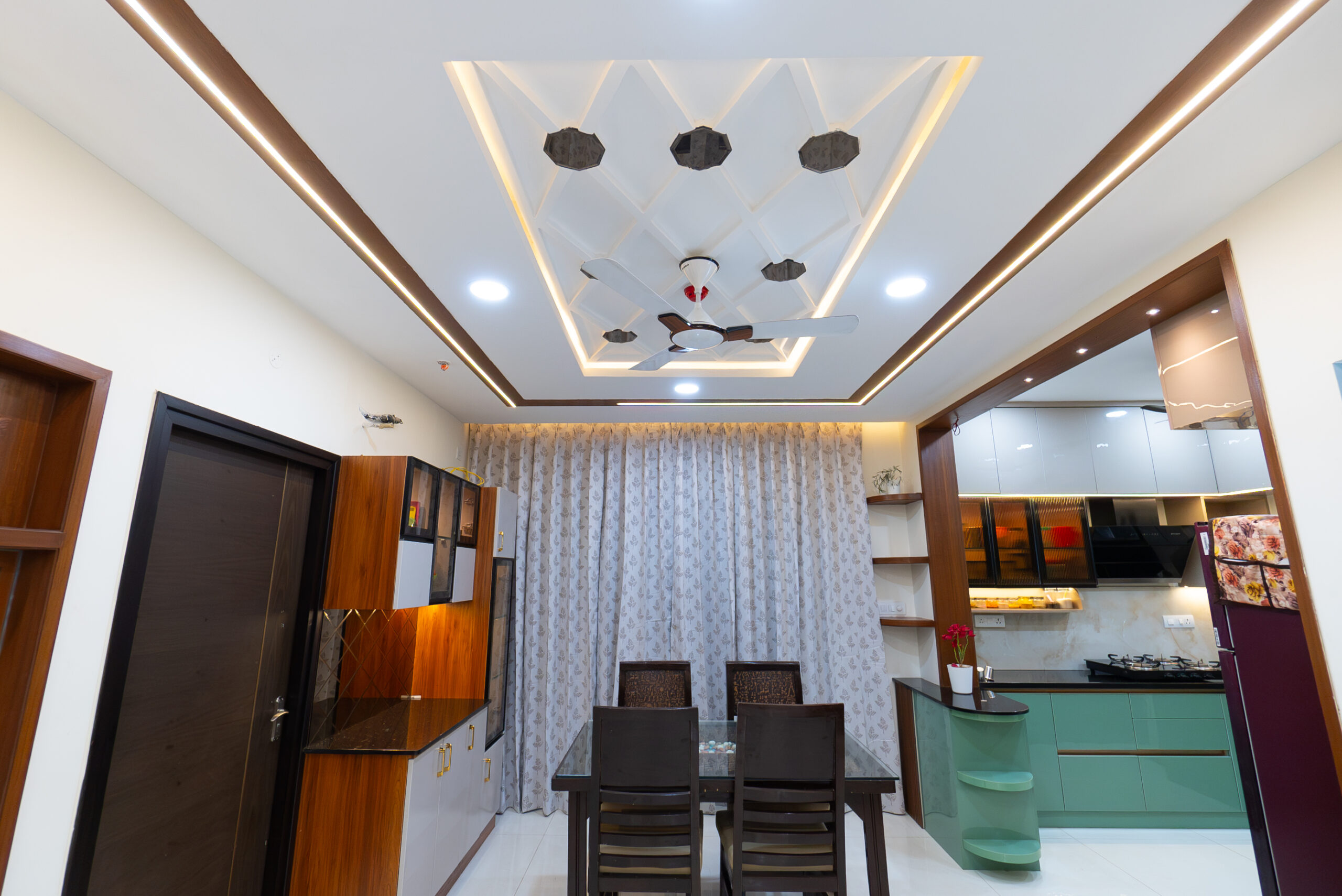 Modern dining and kitchen space designed for home interior renovation in Hyderabad featuring false ceiling lighting, modular cabinets, elegant curtains, and premium finishes.